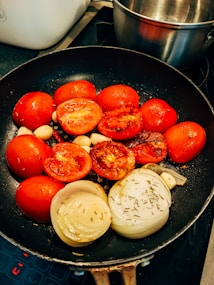 A frying pan contains halved red tomatoes, sliced onions, and garlic cloves being cooked on a stove. The vegetables appear to be slightly charred, suggesting they are being sautéed or grilled. There is a stainless steel pot visible in the background.