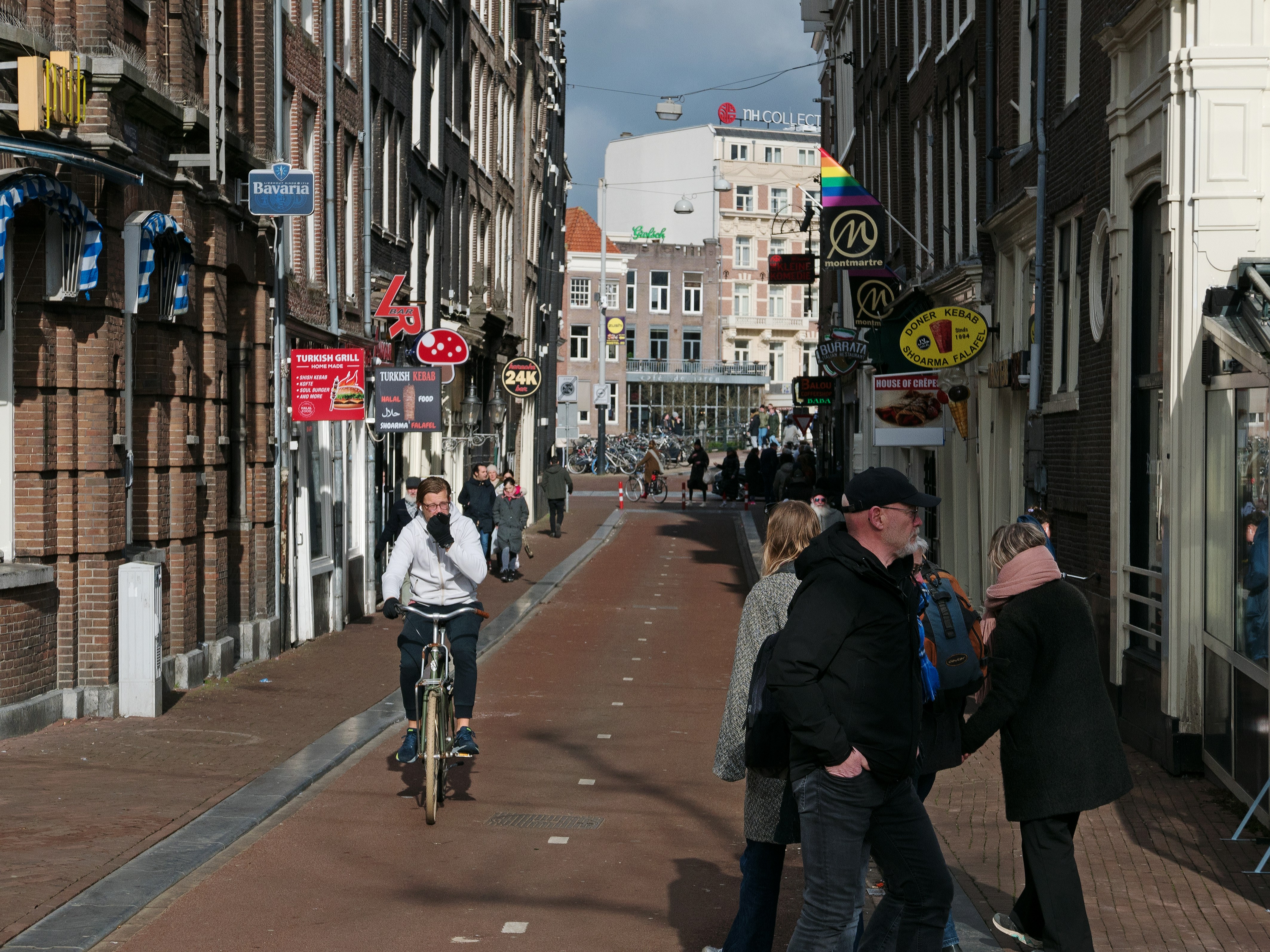 Street view in the alley Halvemaansteeg in Amsterdam old city, on a sunny day with many old house facades and with walking people together, and a biking man alone; photo of Fons Heijnsbroek, Netherlands 19 February 2022. CCO photo in high resolution of mother and kids - Dutch human street photography of sunny city Amsterdam, The Netherlands by Fons Heijnsbroek. Rechtenvrije, gratis foto in hoge resolutie van Amsterdam - mensen lopen en fietsen in de smalle Halvemaansteeg op straat in de zon en schaduw bij het Rembrandtplein; straat-fotografie Nederland, 2022. Op de achtergrond ligt de Amstel.