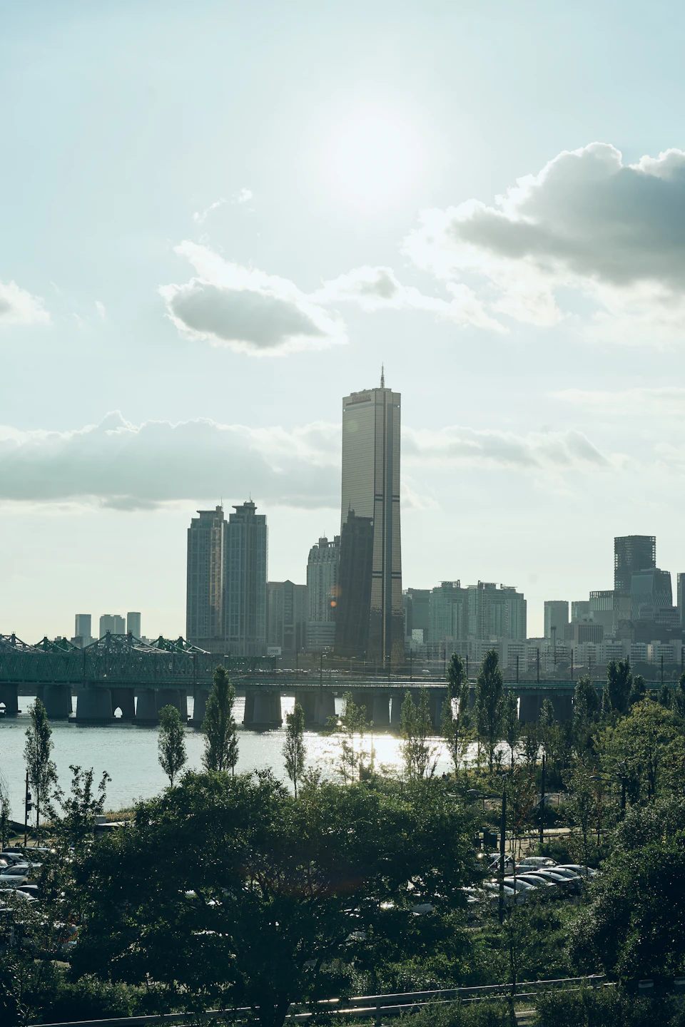 The image depicts a cityscape with a tall, modern skyscraper dominating the skyline. The sun is shining brightly from the top right corner, casting a warm glow over the scene and highlighting the architectural details of the buildings. The city is surrounded by water on three sides, creating a serene backdrop for the urban landscape. In the foreground, there are trees that add a touch of nature to the otherwise concrete environment.
