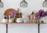 A neatly arranged kitchen shelf displays glass jars filled with flour and oats, a wooden cutting board, a woven placemat, stacks of white bowls, inverted wine glasses, and a vase with vibrant purple flowers. A wooden star decorates the backdrop, and there's also a jar candle on the right.