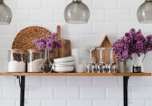 A neatly arranged kitchen shelf displays glass jars filled with flour and oats, a wooden cutting board, a woven placemat, stacks of white bowls, inverted wine glasses, and a vase with vibrant purple flowers. A wooden star decorates the backdrop, and there's also a jar candle on the right.