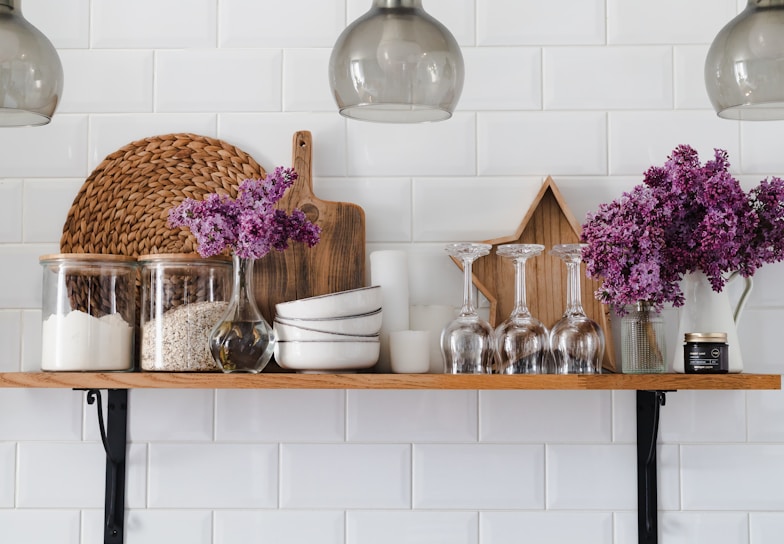 A neatly arranged kitchen shelf displays glass jars filled with flour and oats, a wooden cutting board, a woven placemat, stacks of white bowls, inverted wine glasses, and a vase with vibrant purple flowers. A wooden star decorates the backdrop, and there's also a jar candle on the right.