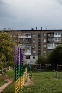 A residential apartment building with a weathered facade stands in the background, surrounded by trees. In the foreground, a playground features colorful climbing structures and monkey bars, with green grass underfoot.