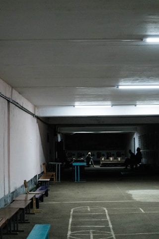A dimly lit indoor space with visible fluorescent lighting on the ceiling. There are several long benches lined up along the walls, and faint outlines of a game is marked on the floor. In the background, there is a group of people sitting around a table with visible numbers 4, 5, and 6, possibly indicating a shooting or archery range.
