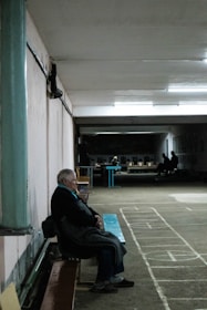 An elderly person sits on a bench in a dimly lit indoor space with marked flooring. In the background, more people are visible, seated and engaged in different activities. The setting suggests a community or recreational area, possibly a basement or hallway with a slightly worn appearance.