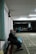 An elderly person sits on a bench in a dimly lit indoor space with marked flooring. In the background, more people are visible, seated and engaged in different activities. The setting suggests a community or recreational area, possibly a basement or hallway with a slightly worn appearance.