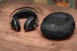 A pair of black, over-ear headphones with silver accents lies on a wooden surface next to a matching black zippered carrying case. The headphones have a sleek, modern design, and the case has a logo embossed on its top.