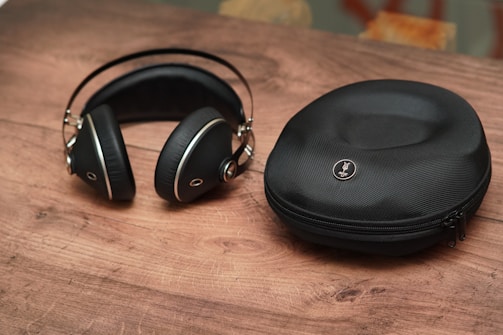 A pair of black, over-ear headphones with silver accents lies on a wooden surface next to a matching black zippered carrying case. The headphones have a sleek, modern design, and the case has a logo embossed on its top.