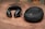 A pair of black, over-ear headphones with silver accents lies on a wooden surface next to a matching black zippered carrying case. The headphones have a sleek, modern design, and the case has a logo embossed on its top.