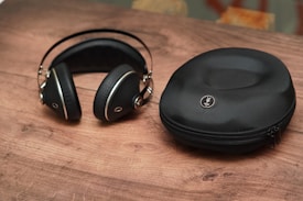 A pair of black, over-ear headphones with silver accents lies on a wooden surface next to a matching black zippered carrying case. The headphones have a sleek, modern design, and the case has a logo embossed on its top.
