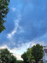 Electric wires stretch across a residential street lined with green trees and a few houses. The sky is filled with clouds, displaying a mix of blue and gray tones.