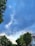 Electric wires stretch across a residential street lined with green trees and a few houses. The sky is filled with clouds, displaying a mix of blue and gray tones.