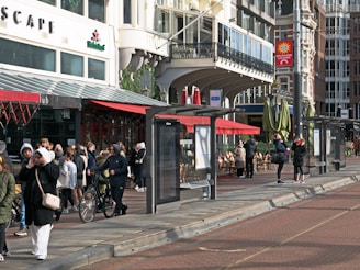 A bustling urban street scene with several people gathering near shops, a cafe, and a tram stop. The architecture is a mix of classic and modern styles with large windows and intricate details. Outdoor seating is visible under red awnings, and people are dressed in winter clothing, some holding bicycles.