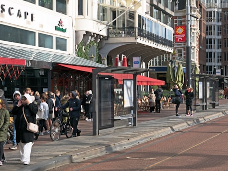 A bustling urban street scene with several people gathering near shops, a cafe, and a tram stop. The architecture is a mix of classic and modern styles with large windows and intricate details. Outdoor seating is visible under red awnings, and people are dressed in winter clothing, some holding bicycles.