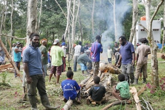 A group of people gathered in a forested area, some standing and others seated around a small fire. The scene includes fallen branches and a smoky atmosphere. Various individuals are engaged in conversation or activities related to the gathering. The background reveals tall, slender trees and a white building with red accents.