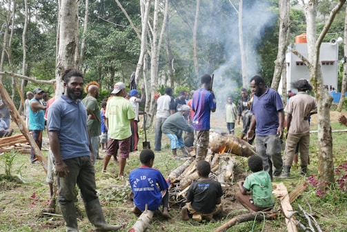 A group of people gathered in a forested area, some standing and others seated around a small fire. The scene includes fallen branches and a smoky atmosphere. Various individuals are engaged in conversation or activities related to the gathering. The background reveals tall, slender trees and a white building with red accents.
