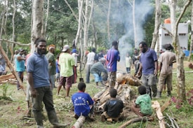 A group of people gathered in a forested area, some standing and others seated around a small fire. The scene includes fallen branches and a smoky atmosphere. Various individuals are engaged in conversation or activities related to the gathering. The background reveals tall, slender trees and a white building with red accents.
