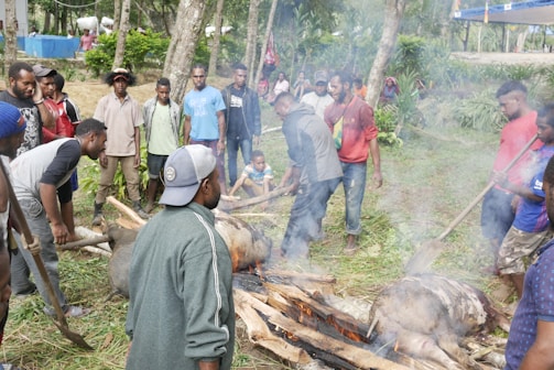 A group of people gathered outdoors around a fire, engaging in a communal cooking process involving large logs and a pig. The setting appears to be rural, surrounded by trees and greenery, with men and a few children participating in or observing the activity.