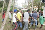 A group of people are gathered outdoors in a wooded area, working together to carry a large object or perhaps an animal. They appear to be focused, collaborating in their efforts. Some are wearing casual clothing, while others are in work attire. The setting seems to be rural, with trees and greenery around.