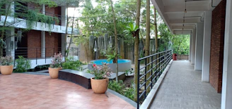 A serene courtyard area with a mix of natural and architectural elements, featuring potted plants on tiled flooring and a raised garden bed enclosing a small circular pool. The space is bordered by modern buildings with red brick walls and large windows, and lush greenery surrounds the area, contributing to a tranquil atmosphere.