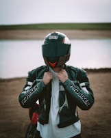 A person stands in front of a body of water wearing a motorcycle helmet and a leather jacket with 'PROBIKER' written on the sleeves. The background features a dirt shore leading to a calm water surface, with a flat expanse of land visible in the distance.