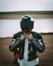 A person stands in front of a body of water wearing a motorcycle helmet and a leather jacket with 'PROBIKER' written on the sleeves. The background features a dirt shore leading to a calm water surface, with a flat expanse of land visible in the distance.