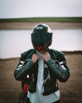 A person stands in front of a body of water wearing a motorcycle helmet and a leather jacket with 'PROBIKER' written on the sleeves. The background features a dirt shore leading to a calm water surface, with a flat expanse of land visible in the distance.