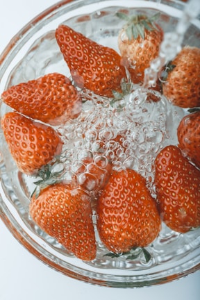 Fresh strawberries submerged in clear water with bubbles forming around them, highlighting their vibrant red color and green leaves. The perspective is top-down, showcasing the texture and details of the fruit.