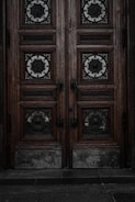 An ornate double door crafted from dark wood features intricate metalwork details. The panels are adorned with circular wrought iron inserts, each with unique, decorative symbols. The door handles are also made of metal, finely detailed in design. The surface of the doors shows signs of wear, lending an aged and historical appearance.