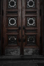 An ornate double door crafted from dark wood features intricate metalwork details. The panels are adorned with circular wrought iron inserts, each with unique, decorative symbols. The door handles are also made of metal, finely detailed in design. The surface of the doors shows signs of wear, lending an aged and historical appearance.
