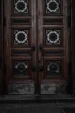 An ornate double door crafted from dark wood features intricate metalwork details. The panels are adorned with circular wrought iron inserts, each with unique, decorative symbols. The door handles are also made of metal, finely detailed in design. The surface of the doors shows signs of wear, lending an aged and historical appearance.