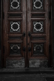 An ornate double door crafted from dark wood features intricate metalwork details. The panels are adorned with circular wrought iron inserts, each with unique, decorative symbols. The door handles are also made of metal, finely detailed in design. The surface of the doors shows signs of wear, lending an aged and historical appearance.