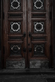 An ornate double door crafted from dark wood features intricate metalwork details. The panels are adorned with circular wrought iron inserts, each with unique, decorative symbols. The door handles are also made of metal, finely detailed in design. The surface of the doors shows signs of wear, lending an aged and historical appearance.