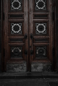 An ornate double door crafted from dark wood features intricate metalwork details. The panels are adorned with circular wrought iron inserts, each with unique, decorative symbols. The door handles are also made of metal, finely detailed in design. The surface of the doors shows signs of wear, lending an aged and historical appearance.