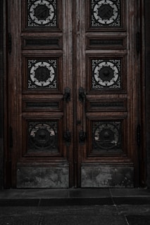 An ornate double door crafted from dark wood features intricate metalwork details. The panels are adorned with circular wrought iron inserts, each with unique, decorative symbols. The door handles are also made of metal, finely detailed in design. The surface of the doors shows signs of wear, lending an aged and historical appearance.