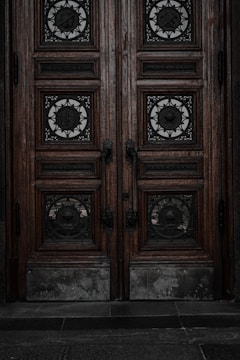 An ornate double door crafted from dark wood features intricate metalwork details. The panels are adorned with circular wrought iron inserts, each with unique, decorative symbols. The door handles are also made of metal, finely detailed in design. The surface of the doors shows signs of wear, lending an aged and historical appearance.