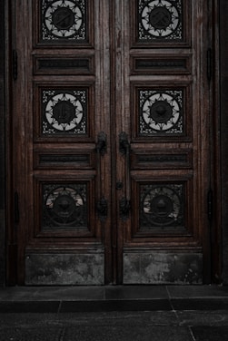 An ornate double door crafted from dark wood features intricate metalwork details. The panels are adorned with circular wrought iron inserts, each with unique, decorative symbols. The door handles are also made of metal, finely detailed in design. The surface of the doors shows signs of wear, lending an aged and historical appearance.