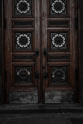 An ornate double door crafted from dark wood features intricate metalwork details. The panels are adorned with circular wrought iron inserts, each with unique, decorative symbols. The door handles are also made of metal, finely detailed in design. The surface of the doors shows signs of wear, lending an aged and historical appearance.