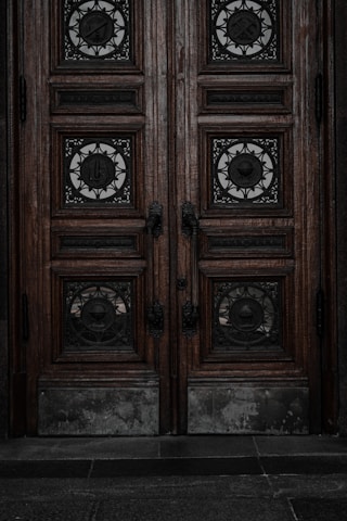 An ornate double door crafted from dark wood features intricate metalwork details. The panels are adorned with circular wrought iron inserts, each with unique, decorative symbols. The door handles are also made of metal, finely detailed in design. The surface of the doors shows signs of wear, lending an aged and historical appearance.