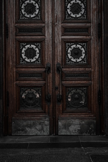 An ornate double door crafted from dark wood features intricate metalwork details. The panels are adorned with circular wrought iron inserts, each with unique, decorative symbols. The door handles are also made of metal, finely detailed in design. The surface of the doors shows signs of wear, lending an aged and historical appearance.