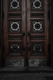 An ornate double door crafted from dark wood features intricate metalwork details. The panels are adorned with circular wrought iron inserts, each with unique, decorative symbols. The door handles are also made of metal, finely detailed in design. The surface of the doors shows signs of wear, lending an aged and historical appearance.