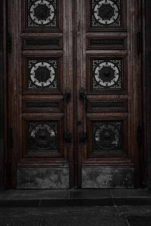 An ornate double door crafted from dark wood features intricate metalwork details. The panels are adorned with circular wrought iron inserts, each with unique, decorative symbols. The door handles are also made of metal, finely detailed in design. The surface of the doors shows signs of wear, lending an aged and historical appearance.