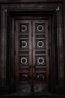 An intricately carved wooden door with ornate geometric and floral patterns. The door is framed by stonework, and the wood appears weathered and aged, giving it a historical feel. The metalwork on the door panels is detailed and decorative.