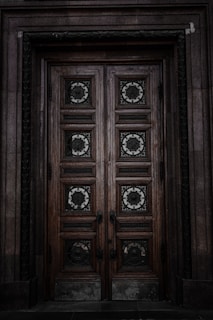 An intricately carved wooden door with ornate geometric and floral patterns. The door is framed by stonework, and the wood appears weathered and aged, giving it a historical feel. The metalwork on the door panels is detailed and decorative.