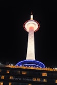 A brightly illuminated tower stands against the night sky, featuring a white cylindrical structure with a circular observation deck lit in blue and red. Below the tower, a building with glowing windows displays the words 'KYOTO TOWER HOTEL.'