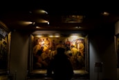 A person is standing in front of a large, framed painting in a dimly lit room. The painting depicts a historical scene with multiple figures wearing period clothing, including hats and traditional attire. The focus is on the art piece, highlighted by overhead lights, while the rest of the room remains in shadow.