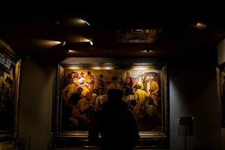A person is standing in front of a large, framed painting in a dimly lit room. The painting depicts a historical scene with multiple figures wearing period clothing, including hats and traditional attire. The focus is on the art piece, highlighted by overhead lights, while the rest of the room remains in shadow.