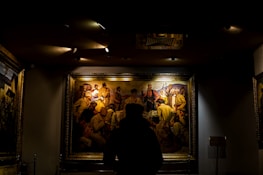 A person is standing in front of a large, framed painting in a dimly lit room. The painting depicts a historical scene with multiple figures wearing period clothing, including hats and traditional attire. The focus is on the art piece, highlighted by overhead lights, while the rest of the room remains in shadow.