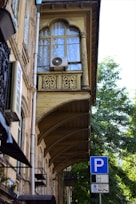 A historic building with ornate architectural details, including decorative arches and intricate woodwork on balconies. An air conditioner unit is mounted on a window, which is covered with translucent curtains. A blue parking sign is visible in the foreground, set against a background of leafy green trees and a clear sky.