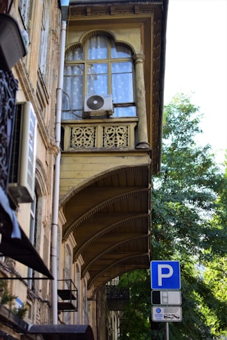 A historic building with ornate architectural details, including decorative arches and intricate woodwork on balconies. An air conditioner unit is mounted on a window, which is covered with translucent curtains. A blue parking sign is visible in the foreground, set against a background of leafy green trees and a clear sky.